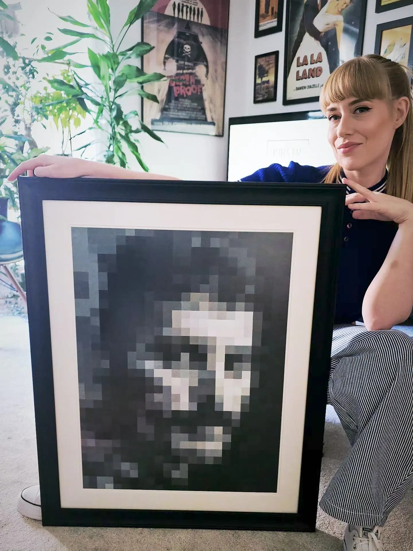 A woman with bangs and a ponytail kneels, holding up a framed piece of pixel art that depicts a face.